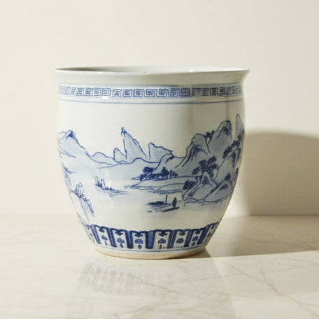 A large ceramic planter pot with a white glaze and blue hand-painted landscape design.