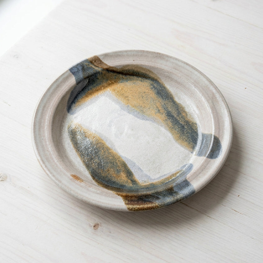 A large round ceramic plate with an abstract painted design in gray, gold, and brown.