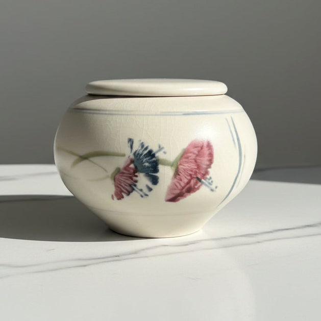 A small cream-colored ceramic lidded jar decorated with pink and dark blue flowers.