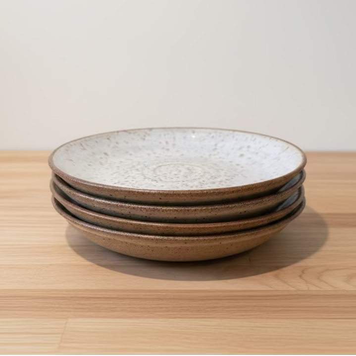 A stack of four round ceramic plates with a speckled white interior and a natural base.