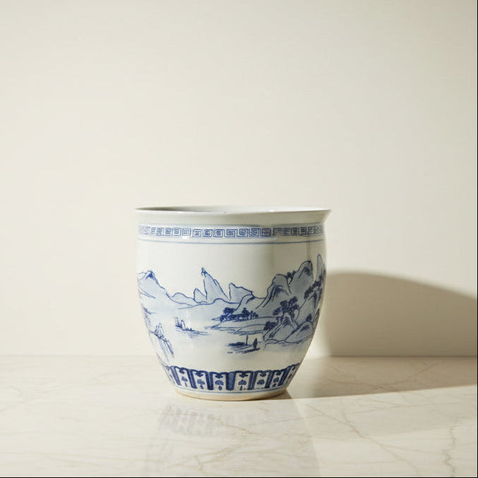 A large ceramic planter pot with a white glaze and blue hand-painted landscape design.