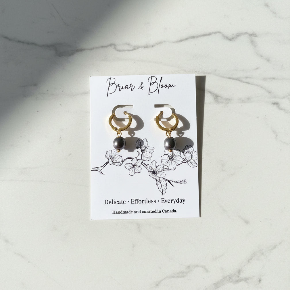 A pair of gold hoop earrings with dark blue grey pearl drops.