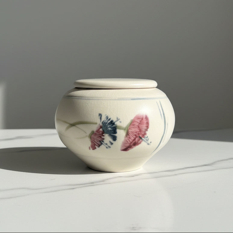 A small cream-colored ceramic lidded jar decorated with pink and dark blue flowers.