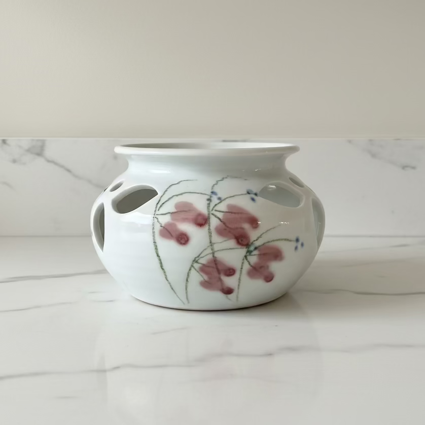 A white ceramic planter pot with side drainage holes, decorated with a hand-painted floral pattern of dusty pink blossoms and green stems.