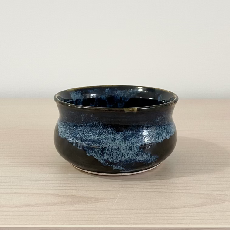 A hand holds a small ceramic bowl with a glossy dark blue and black glaze featuring a subtle speckled pattern.