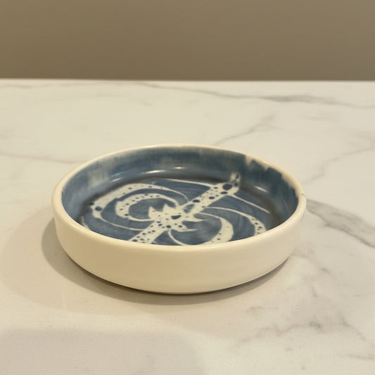 A handmade ceramic dish held in a hand, featuring a blue glazed interior pattern and a solid cream-colored exterior.