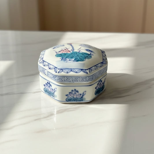 A small  lidded ceramic jar with a painted landscape design in blue, green, and pink on a white base