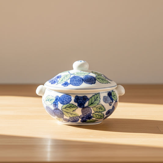 A small white ceramic lidded bowl decorated with blue berries and green leaves.