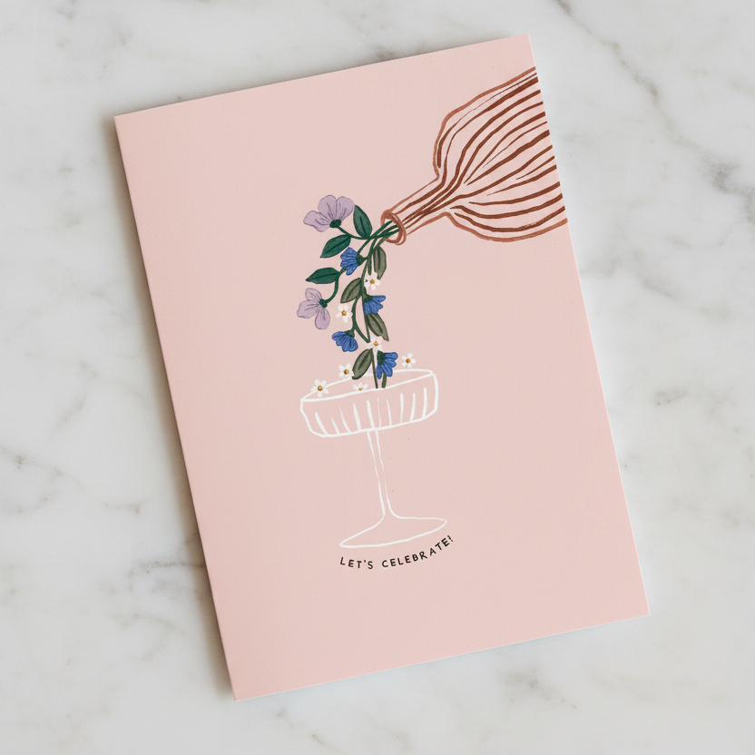 A pink greeting card with a bottle and glass reading " Let's Celebrate"