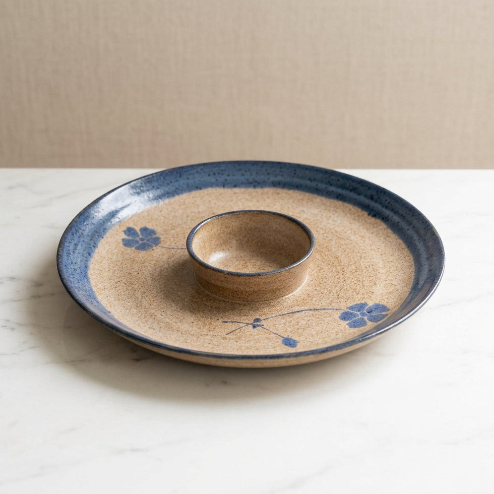 A round ceramic serving platter with a central dipping bowl, featuring a speckled beige glaze, a dark blue rim, and blue floral motifs.