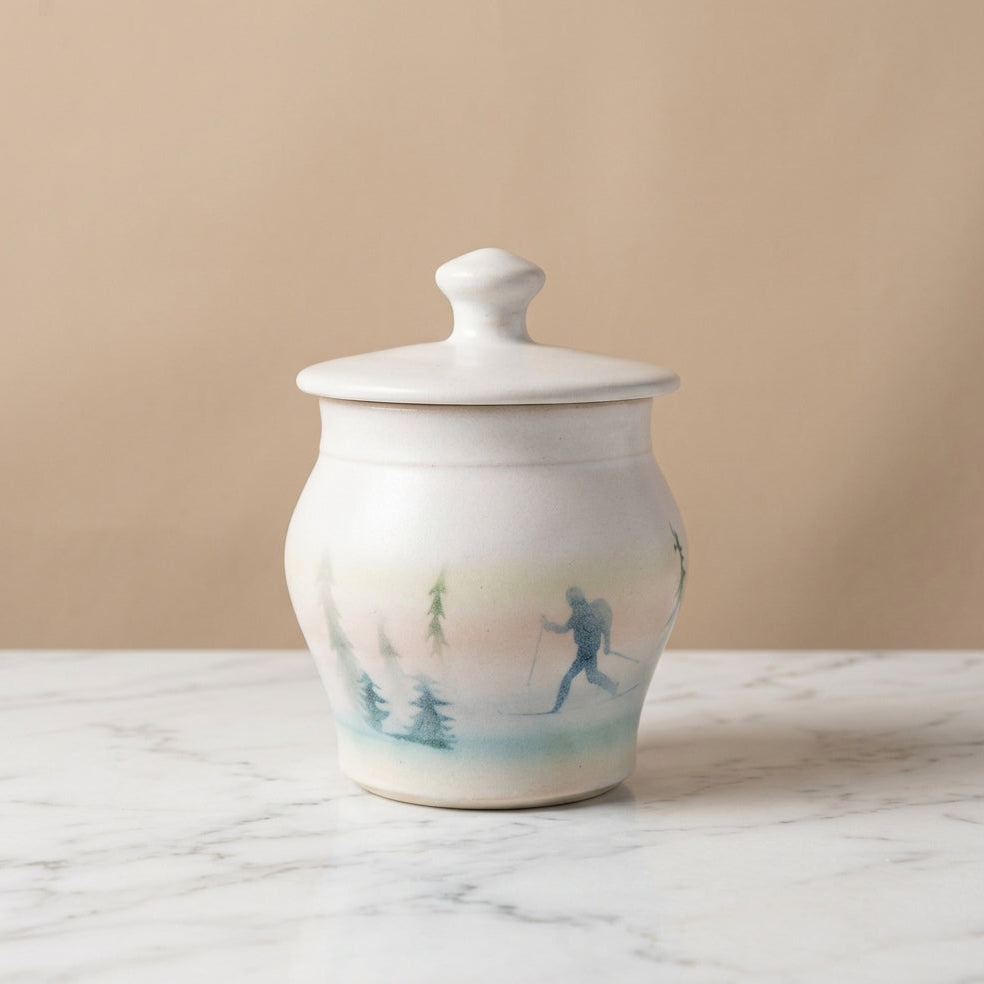 A white ceramic lidded jar with a hand-painted winter landscape design on the front, featuring pine trees and a skier in blue and green tones.