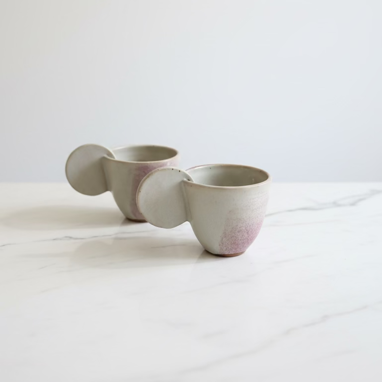 Two handmade ceramic mugs with integrated handles and lids, featuring a dusty rose pink wash at the base.